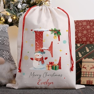 May include: A beige Christmas gift bag with a red drawstring. The bag features a large red letter "E" with a snowman, snowflakes, and the text "Merry Christmas Evelyn". A Christmas tree and wrapped gifts are in the background.