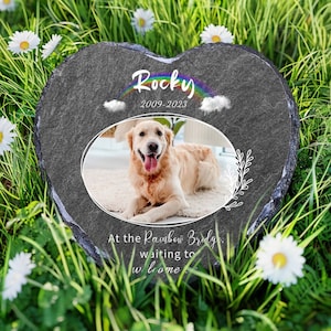 May include: A heart-shaped gray stone memorial with a photo of a golden retriever, the name "Rocky" and the dates "2009-2023" inscribed on it. The text "At the Rainbow Bridge, waiting to welcome you home" is also inscribed on the stone.