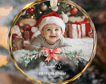 May include: A round, clear glass Christmas ornament with a gold trim. The ornament features a photo of a baby wearing a Santa hat, surrounded by greenery and a red bow. The text "Sarah's First Christmas 2024" is printed on the ornament.