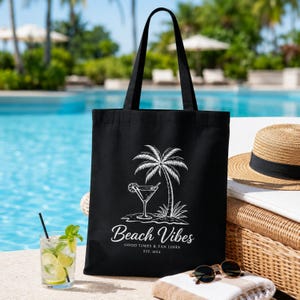 May include: A black canvas tote bag featuring a white graphic print of a palm tree and a cocktail glass. The text reads Beach Vibes, GOOD TIMES & TAN LINES, EST. 2024. The bag has two long black shoulder straps and a rectangular body shape.