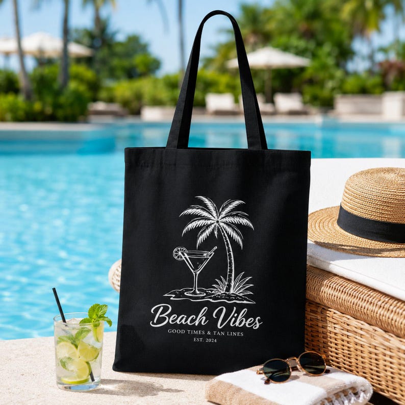 May include: A black canvas tote bag featuring a white graphic print of a palm tree and a cocktail glass. The text reads Beach Vibes, GOOD TIMES & TAN LINES, EST. 2024. The bag has two long black shoulder straps and a rectangular body shape.