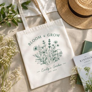 May include: A cream-colored canvas tote bag featuring a dark green botanical illustration of wildflowers. The text reads BLOOM & GROW, Emily's Garden, and EST. 2024. The bag has two long shoulder straps and is made of a durable, woven fabric material.