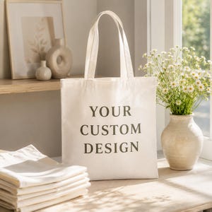 May include: A cream-colored canvas tote bag featuring the dark gray text YOUR CUSTOM DESIGN printed on the front. The bag has two long shoulder straps and is positioned upright next to a stack of folded fabric and a vase of white flowers.