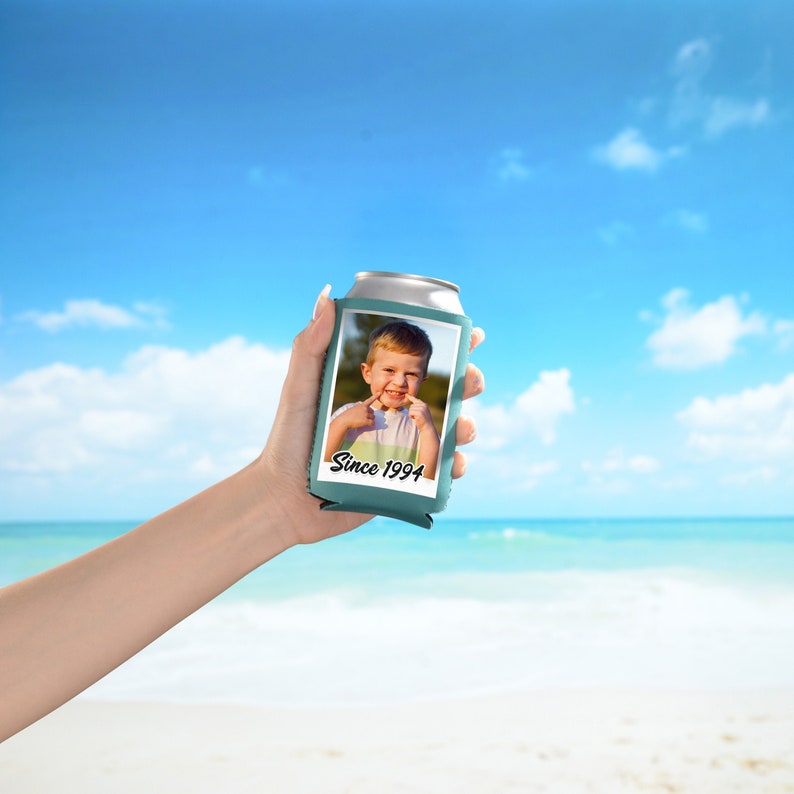 May include: A teal blue can cooler with a photo of a young boy smiling and pointing to his teeth. The text "Since 1994" is printed on the cooler.