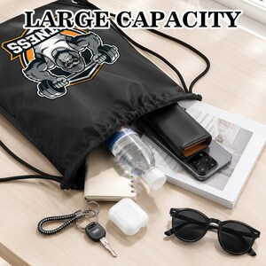 May include: A black drawstring backpack featuring a graphic of a gorilla lifting a barbell with the text LARGE CAPACITY. The bag contains a clear water bottle, a black wallet, a smartphone, a notebook, white earbuds, a key, and black sunglasses.