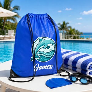 May include: A bright blue drawstring backpack featuring a circular graphic of a teal dolphin jumping over stylized waves. The name James is printed in white script below the dolphin. The bag has black cord straps and reinforced corners.