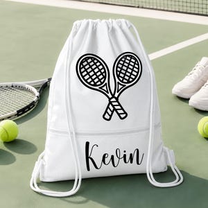 May include: White drawstring sports bag with a black graphic of two crossed tennis rackets. The bag has the name "Kevin" printed in black script. A tennis racket and ball are visible in the background.