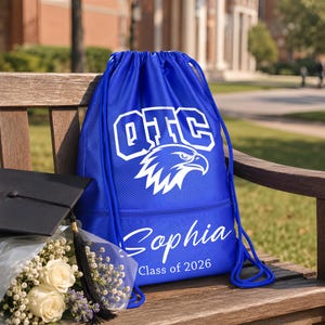 May include: A royal blue drawstring backpack with white text and a graphic of an eagle. The backpack has the text "OTC", "Sophia", and "Class of 2026". A black graduation cap and a bouquet of white roses are also in the image.