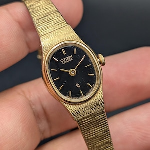 Könnte beinhalten: Eine goldfarbene Citizen Quartz Armbanduhr mit einem ovalen schwarzen Zifferblatt und goldenen Zeigern. Die Uhr hat ein strukturiertes, goldfarbenes Metallarmband mit horizontalen Linien. Das Zifferblatt zeigt den Text Citizen Quartz.