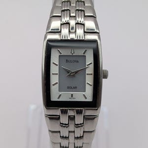May include: A rectangular Bulova solar watch with a silver-toned dial, black border, and silver hands. The watch features a metallic link bracelet with a polished finish. The dial displays the Bulova logo, the word SOLAR, and the model code P4L004.