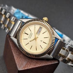 May include: A Citizen Quartz watch with a gold-toned face and silver and gold-toned band. The watch face displays the day and date. The watch is displayed on a brown wooden block against a dark background.
