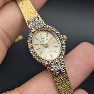 Può includere: Un orologio al quarzo Waltham color oro con quadrante ovale, fondo argento e indici dorati. La lunetta è decorata con piccole pietre trasparenti e due motivi a farfalla color argento in alto e in basso. Presenta un cinturino dorato testurizzato.