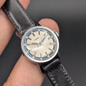 Könnte beinhalten: Eine Vintage-Armbanduhr von Timex mit einem runden, silberfarbenen Zifferblatt, schwarzen Stundenmarkierungen und Zeigern. Die Uhr hat ein poliertes, silberfarbenes Metallgehäuse und ein schwarzes Lederarmband mit sichtbaren Nähten.