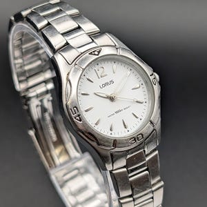 Pode incluir: Um relógio de pulso Lorus prateado com mostrador branco, marcadores de hora e ponteiros prateados. O relógio possui uma pulseira de elos de aço inoxidável e uma luneta giratória com os números 15, 30 e 45 gravados. O mostrador exibe o logo Lorus.