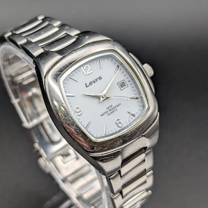 May include: A silver-tone analogue wristwatch with a square-shaped case and a matching metal link bracelet. The white dial features the Levi's logo, silver hands, a date window at the three o'clock position, and the text 3ATM WATER RESISTANT QUARTZ.