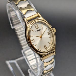 Könnte beinhalten: Eine Timex Armbanduhr mit rundem goldfarbenem Gehäuse und silberfarbenem Zifferblatt mit goldfarbenen Ziffern auf den 3-, 6-, 9- und 12-Uhr-Positionen. Die Uhr hat ein zweifarbiges Metallgliederarmband mit abwechselnd gold- und silberfarbenen Segmenten.