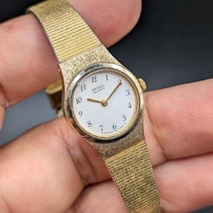 May include: A gold-tone Seiko Quartz wristwatch featuring a white round dial with black Arabic numerals, gold-tone hands, and a textured gold-tone metal link bracelet. The watch face displays the text Seiko Quartz and Japan 2Y00-0260.