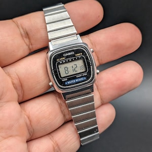 May include: A silver Casio digital watch with a rectangular face and a metal strap. The watch shows the time 8:12 and the date 18. The watch has the words "CASIO", "ALARM", "TIMER", "MODE", "START-STOP", and "WATER RESIST" printed on the face.