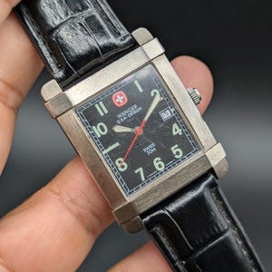 May include: A rectangular silver-tone wristwatch with a black dial, featuring the text Wenger S.A.K. Design, Swiss 30M, and T-Swiss Made T. The watch has a black leather strap with a crocodile-embossed texture, white Arabic numerals, and a red second hand.