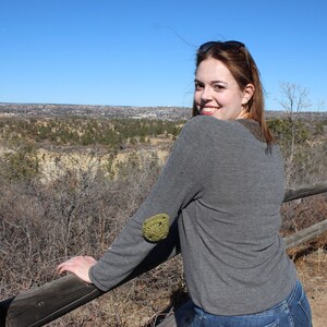Könnte beinhalten: Eine Frau trägt einen grauen Pullover mit einem grünen gehäkelten Flecken am Ellbogen und steht vor einem Holzzaun mit Blick auf eine ferne Stadt.