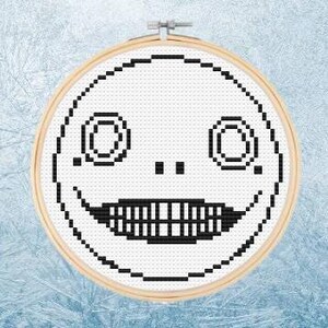 May include: Black and white cross-stitch pattern of a smiling face with large eyes and teeth. The face is stitched on a white fabric and framed in a wooden hoop.