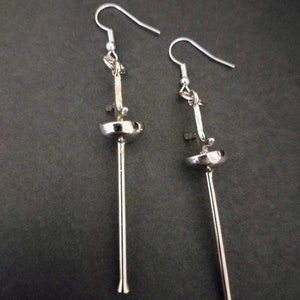 May include: A pair of silver dangle earrings with a long, thin, straight rod and a small, round, silver disc at the top. The earrings have a simple, minimalist design.