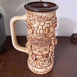 May include: A tall ceramic beer mug with a beige finish and a dark brown rim. The exterior features a relief design of a small house surrounded by foliage and a brick-like pattern. The base includes the Cyrillic text "Ужгород" in raised lettering.