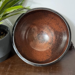 May include: A round ceramic bowl featuring a deep, glossy interior with a mottled copper and black finish. The exterior rim is smooth and dark, transitioning into a textured, dark brown surface. The bowl has a wide, open circular opening.