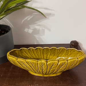 May include: An elongated, oval-shaped ceramic bowl in a deep olive green colour. The exterior features a raised, moulded leaf pattern with defined veins, while the rim has a scalloped edge. The bowl sits on a small, oval-shaped pedestal base.