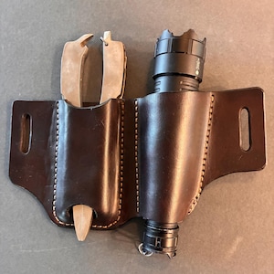 May include: A dark brown leather multi-tool sheath with a flashlight and a multi-tool. The sheath has two pockets, one for the flashlight and one for the multi-tool. The sheath is stitched with light brown thread.