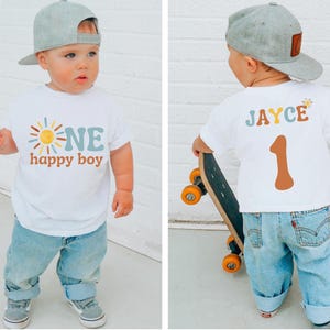 Puede incluir: Una camiseta blanca con las palabras "ONE happy boy" y un gráfico de sol. El niño lleva vaqueros azul claro, zapatos grises y una gorra de béisbol gris. La parte trasera de la camiseta tiene el nombre "JAYCE" y el número "1".