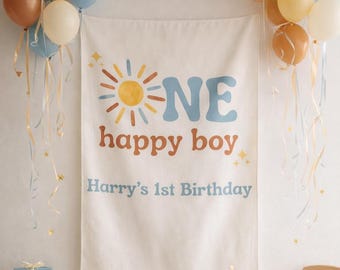 May include: A white fabric banner featuring a yellow sun graphic, the text "ONE happy boy" in blue and brown, and "Harry's 1st Birthday" in blue. The banner is surrounded by clusters of blue, tan, and white balloons with long curling ribbons.