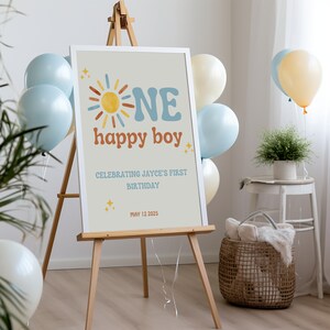 Puede incluir: Un letrero enmarcado sobre un caballete de madera con las palabras "ONE happy boy" y "Celebrating Jayce's First Birthday" en una fuente retro. El letrero est&aacute; decorado con un sol y estrellas. Globos en tonos azules y crema rodean el letrero.