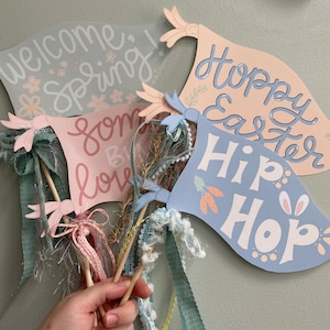 May include: Four pastel blue and pink paper banners with spring and Easter themes. The banners read "Welcome Spring!" "Some Bunny Loves You" "Happy Easter" and "Hip Hop".