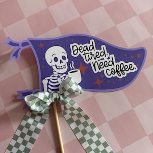 May include: A purple flag-shaped decoration with the text "Dead, tired, Need coffee." A white skeleton is holding a coffee cup. The decoration has a green and white checkered bow and a wooden stick.