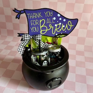 May include: A black cauldron-shaped container filled with Halloween-themed treats. A purple flag with the words "Thank You For All You Brew" is attached. The container includes a jar with ghost designs, a can, and a snack bag.