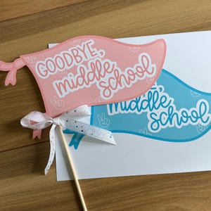 Può includere: Due cartelli di carta rosa e blu con il testo "Goodbye, middle school" e "Middle school". I cartelli sono attaccati a un bastone di legno con un nastro bianco.