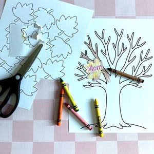 May include: A craft project with a leaf-themed drawing and cutouts. A tree drawing has a leaf with the word "Mom" on it. Crayons in orange, green, pink, and yellow are scattered around the drawing. Scissors and leaf cutouts are also present.