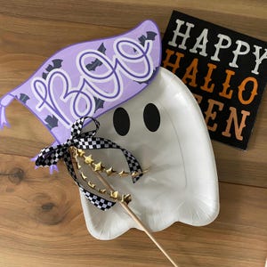 May include: A white ghost-shaped paper plate with black eyes and a purple "Boo" sign with bats on it. A black and white checkered ribbon with gold stars is attached to a wooden stick.