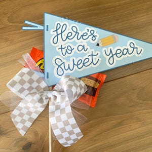 Op de afbeelding: Een blauwe vlag met de tekst "Here's to a sweet year" en een gele potlood-illustratie. Een geruite strik met een tule-overlay is bevestigd aan de vlag.