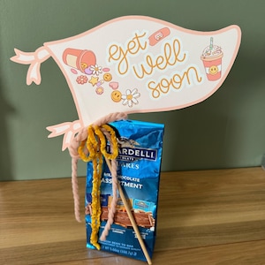 以下が含まれることがあります： ピンクと白の「get well soon」のサインに、漫画のばんそうこう、薬瓶、アイスコーヒーのカップが描かれています。サインは、青と白のチョコレートバーのパッケージに、黄色とピンクのパイプクリーナーで取り付けられています。