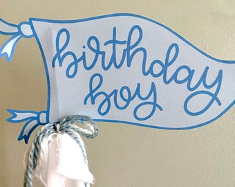 May include: A blue and white banner with the text "birthday boy" in blue cursive writing. The banner is attached to a blue and white rope.