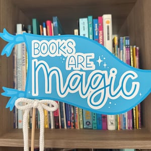 May include: A light blue pennant with the words "BOOKS ARE MAGIC" in white script and block letters. The pennant is attached to a wooden dowel with a white rope bow. A bookshelf filled with books is in the background.