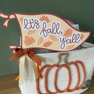 Puede incluir: Una cesta de mimbre blanca con un diseño de calabaza marrón. Una pancarta con el texto "It's fall, y'all" está unida a la cesta con una cinta marrón.