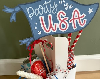 May include: A patriotic party favor basket with a blue banner that reads "Party in the USA." The basket contains a soda can, red and white striped straws, and a star-shaped decoration. The color scheme is red, white, and blue.