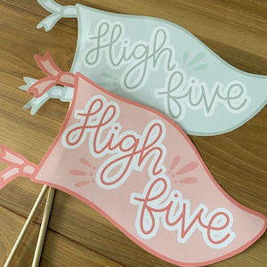 Puede incluir: Dos banderas de fiesta de papel con las palabras "high five" escritas en ellas. Una bandera es rosa con letras blancas y la otra es azul claro con letras grises.
