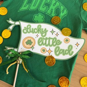 Può includere: Uno striscione bianco con il testo "lucky little lad" in lettere verdi e arancioni. Lo striscione è decorato con trifogli e stelle. Lo striscione è attaccato a un bastone verde e dorato con un campanello.