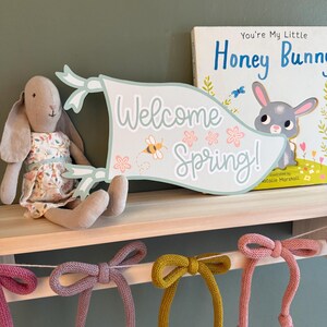 May include: A light blue and green banner with the text "Welcome Spring!" and a bee illustration. The banner is hanging on a wooden shelf with a stuffed bunny and a book titled "You're My Little Honey Bunny" in the background.