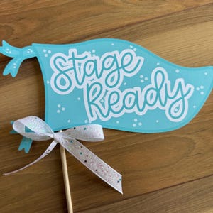 May include: A light blue flag-shaped decoration with the words "Stage Ready" in white script. The flag has a white ribbon with glitter and is attached to a wooden stick. The background is a wooden surface.