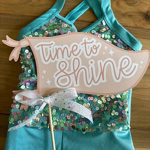 May include: A teal sequined leotard with a pink and white sign that says "Time to Shine". The sign is attached to a stick with a white and pink ribbon.
